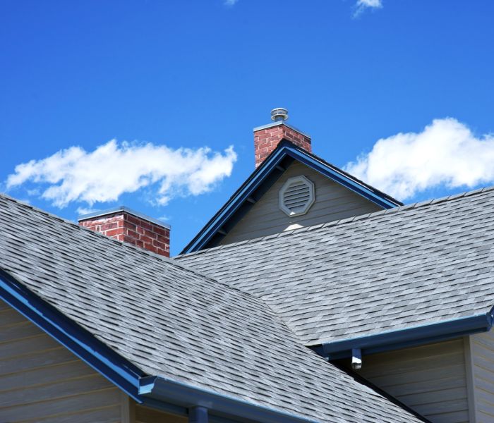 A close-up of a residential roof featuring gray asphalt shingles, a chimney with red bricks, and blue-painted trim.